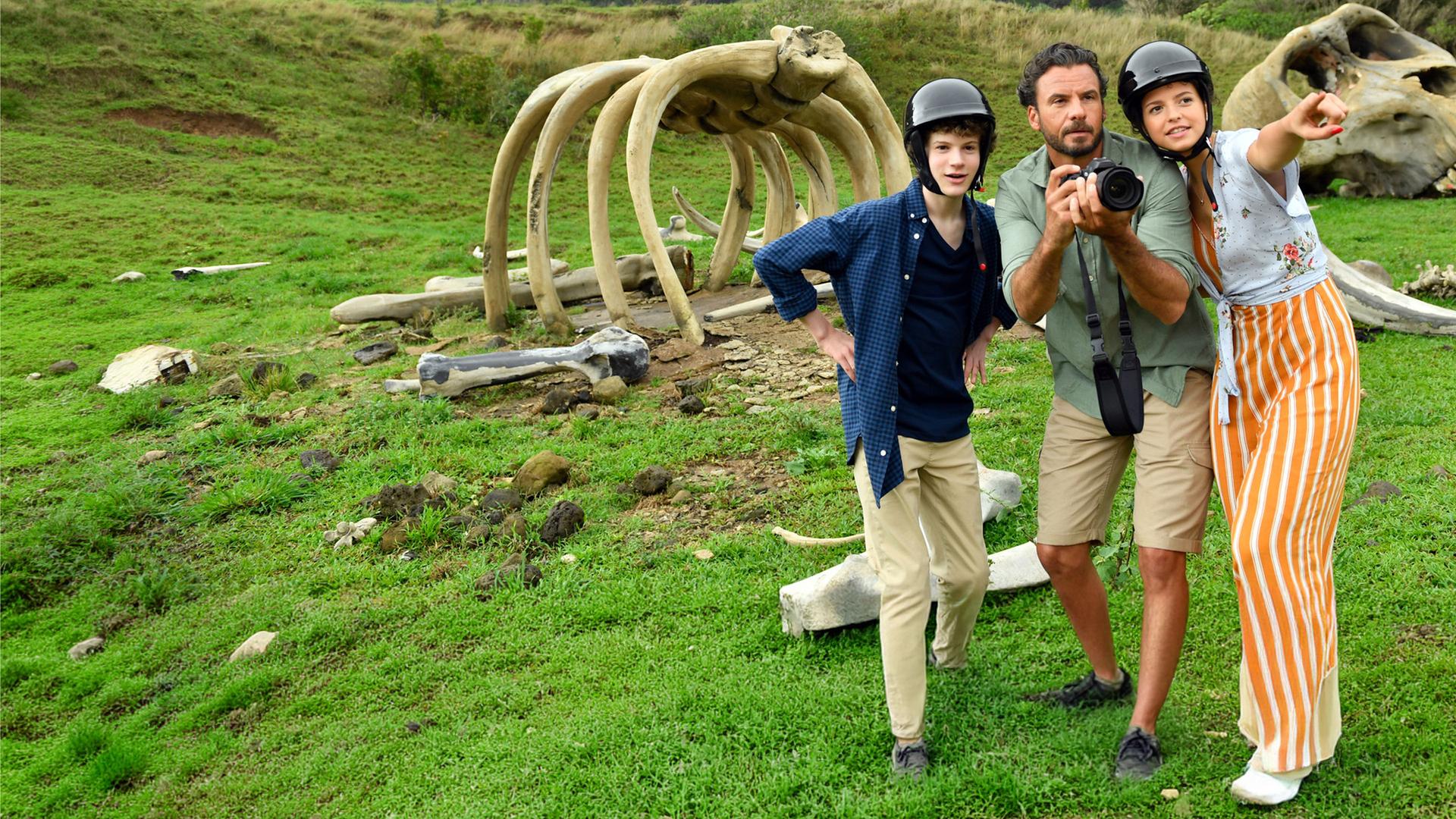 "Das Traumschiff: Hawaii": Jon (Stephan Luca) steht zwischen Moritz (Nico Ramon Kleemann) und Lotte (Nina Kaiser) und hält einen Fotoapparat in der Hand. Die beiden Kinder tragen Helme, Lotte zeigt mit dem Finger auf etwas in der Ferne. Hinter ihnen liegen, vor einem Felsmassiv, große Knochen.