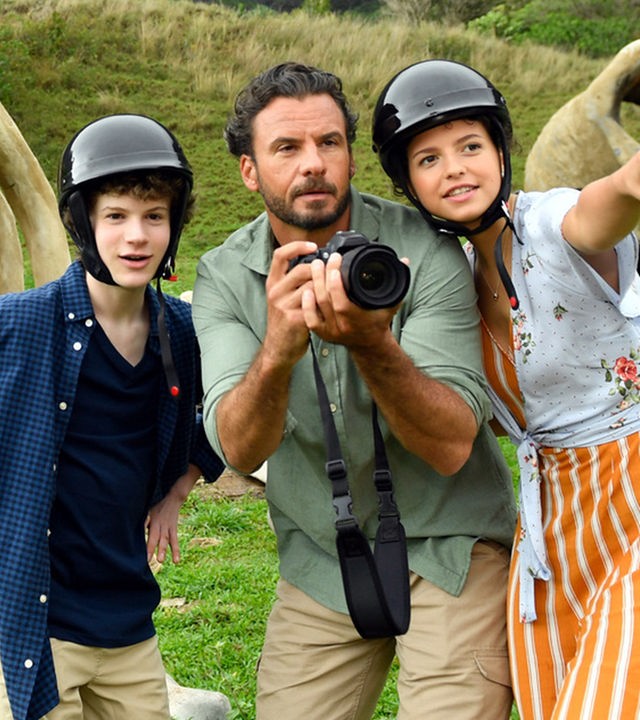 "Das Traumschiff: Hawaii": Jon (Stephan Luca) steht zwischen Moritz (Nico Ramon Kleemann) und Lotte (Nina Kaiser) und hält einen Fotoapparat in der Hand. Die beiden Kinder tragen Helme, Lotte zeigt mit dem Finger auf etwas in der Ferne. Hinter ihnen liegen, vor einem Felsmassiv, große Knochen.