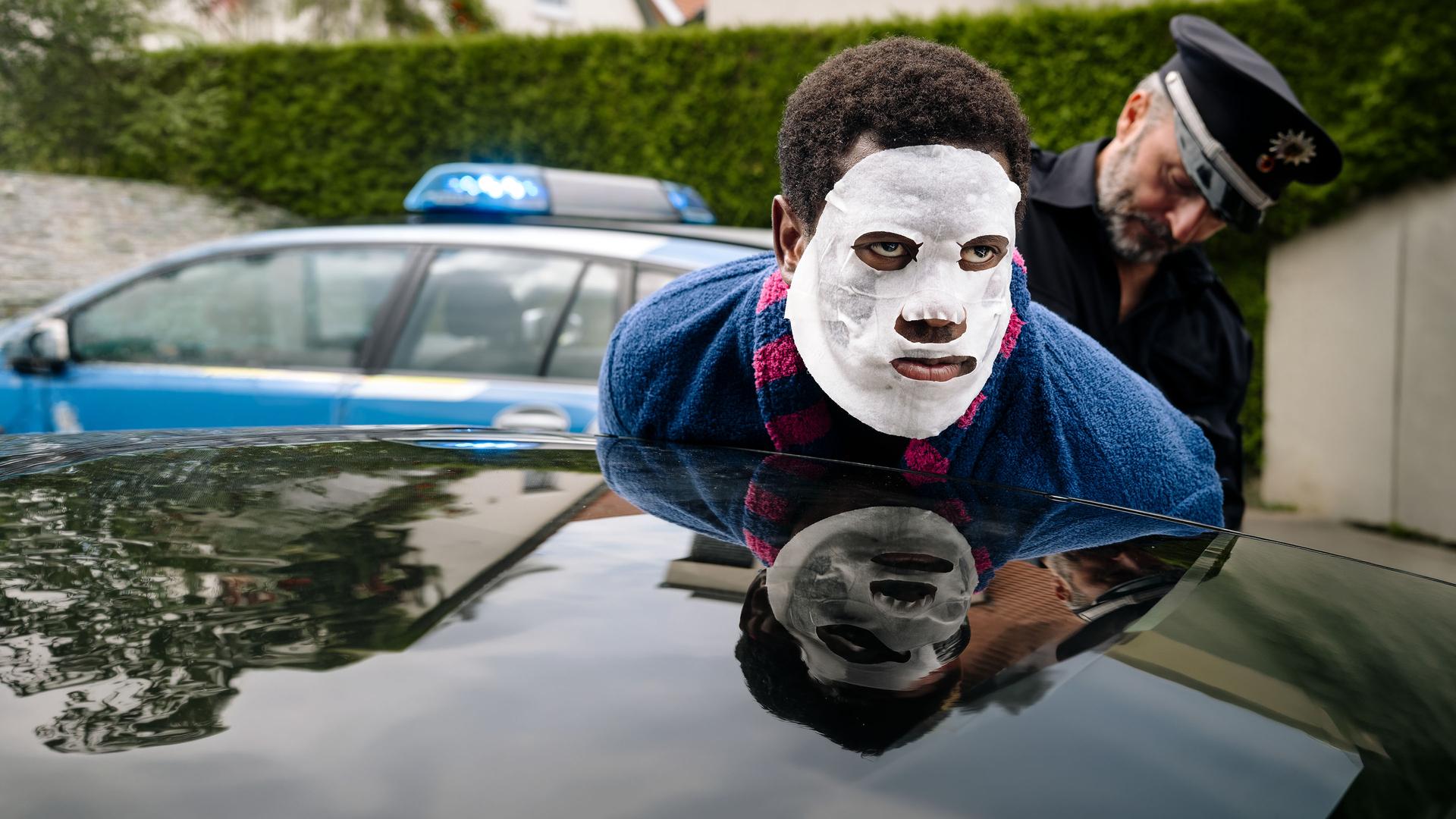 "I don't work here - Hecke": Dawit (Akeem van Flodrop) wird von Polizist Jakob (Robert Schupp) festgenommen. Während Jakob ihn in Polizeiuniform an der Autotür fixiert, lehnt Dawit seinen Oberkörper an ein dunkles Autodach und trägt eine weiße Tuchmaske und einen blauen Bademantel mit pinken Streifen.