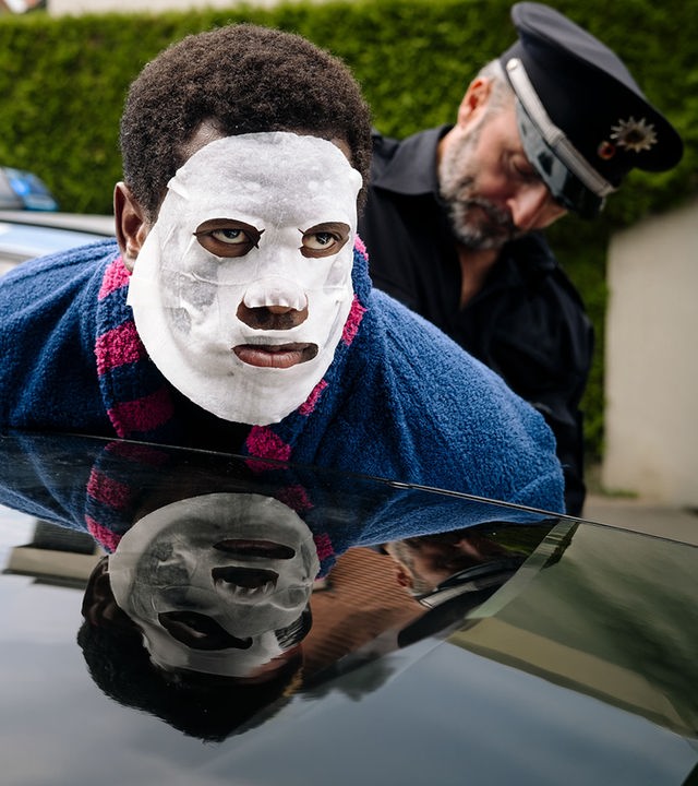 "I don't work here - Hecke": Dawit (Akeem van Flodrop) wird von Polizist Jakob (Robert Schupp) festgenommen. Während Jakob ihn in Polizeiuniform an der Autotür fixiert, lehnt Dawit seinen Oberkörper an ein dunkles Autodach und trägt eine weiße Tuchmaske und einen blauen Bademantel mit pinken Streifen.