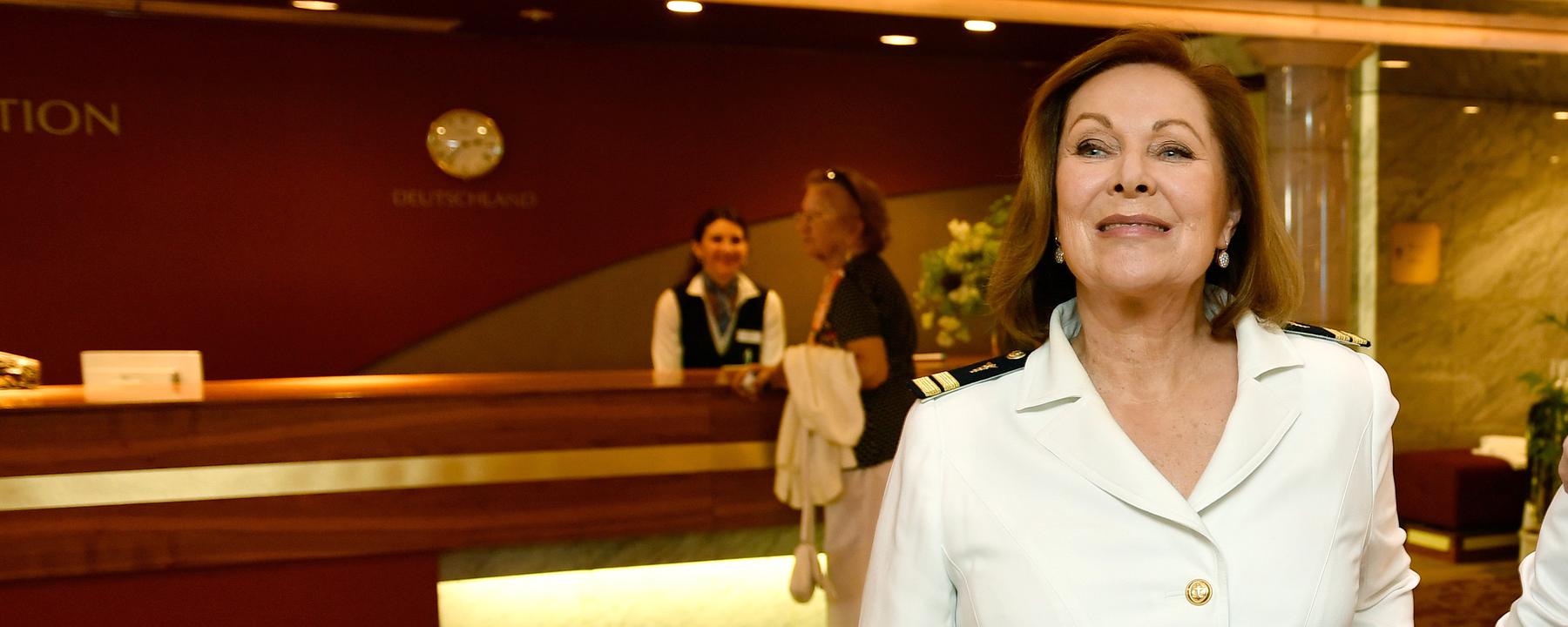 Beatrice (Heide Keller) steht am inneren Treppenaufgang im Schiff. Im Hintergrund sieht man die Rezeption des Schiffs.