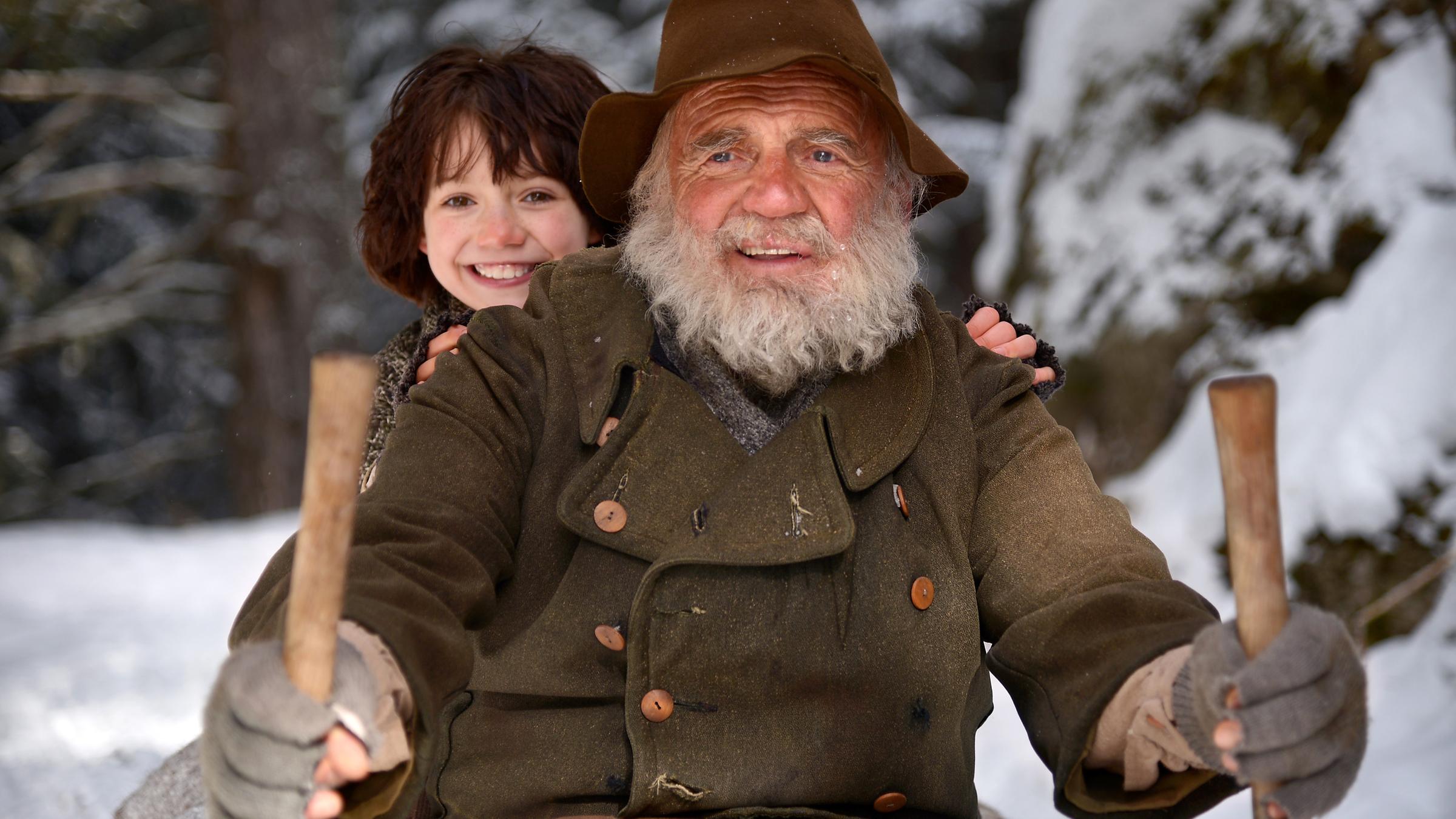 Der Almöhi (Bruno Ganz) und Heidi (Anuk Steffen) rodeln mit ihrem Schlitten durch die verschneiten Berge ins Dorf. Heidi sitzt hinter ihrem Großvater und schaut ihm verschmitzt lachend, mit roter Nasenspitze über die Schulter. Auch der Almöhi hat ein breites Grinsen im Gesicht.