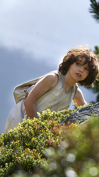 Heidi klettert einen großen, grün bewachsenen Felsen hoch und schaut neugierig über die Kuppe. Vor ihr liegt versteckt eine Ziege im Gras und über ihr ist der blaue Himmel zu sehen.