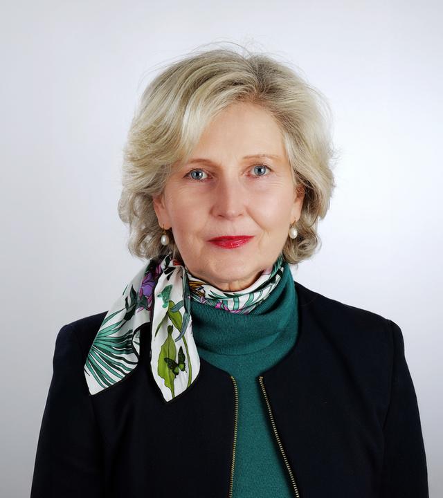 Porträtfoto einer Frau mittleren Alters mit kurzen graumelilerten Haaren, die ein schwarze Jacke über einem Rollkragenpullover und kurzen Schal trägt.