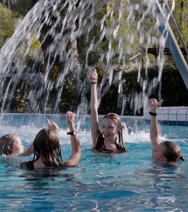 Das Bild zeigt eine fröhliche Szene in einem Außenpool, wahrscheinlich in einem Thermalbad. Im Vordergrund sind vier Kinder zu sehen, die im Wasser spielen. Sie haben nasse Haare und tragen Badebekleidung. Die Kinder zeigen mit ihren Händen nach oben, offenbar erfreut über das Wasser, das aus einer etwa zwei Meter hohen Wasserrampe herabfließt.  Im Hintergrund ist ein älterer Mann zu erkennen, der entspannt im Wasser sitzt. Der Pool ist von einer grünen Hecke umgeben, und im Hintergrund sind Bäume und weitere Freizeitbereiche sichtbar. Der Himmel scheint sonnig, was auf angenehmes Wetter hindeutet. Die gesamte Szene vermittelt ein Gefühl von Freude, Erholung und Freizeitaktivität.