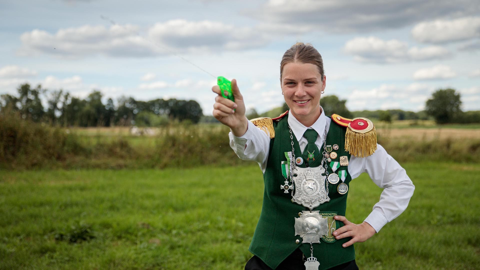 Anneke steht mit einer Wasserspritzpistole in der Hand auf einem Feld und drückt ab.