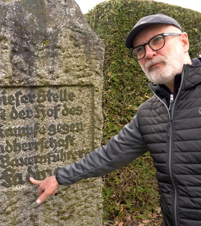 Ein Mann zeigt auf einen Namen auf einem verwitterten Gedenkstein