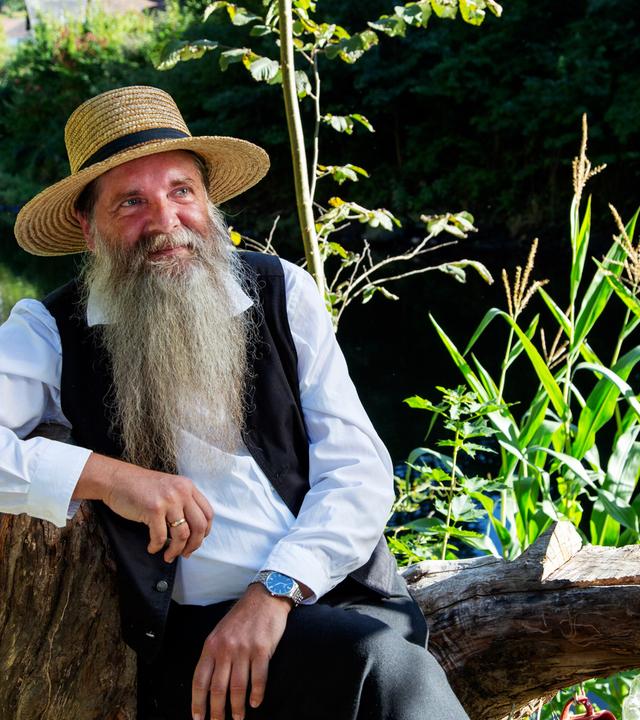 Ein Mann mit langem Bart und Strohhut sitzt auf einem Baumstamm im Freien.