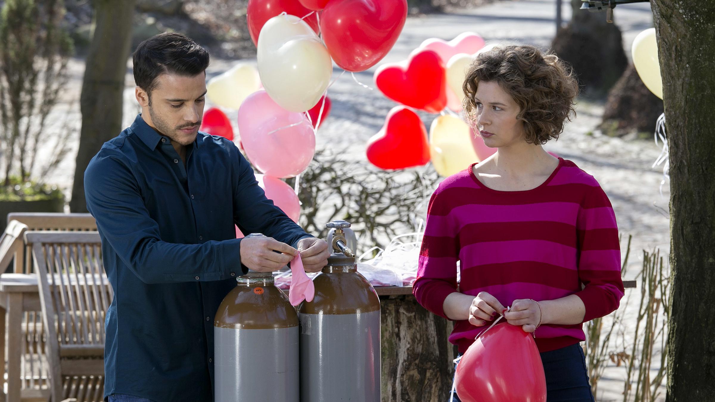 Kathi (Anna Maria Sturm) und Theo (Kostja Ullmann) beim Füllen der Herz-Luftballons. Kathi schaut Theo böse an.