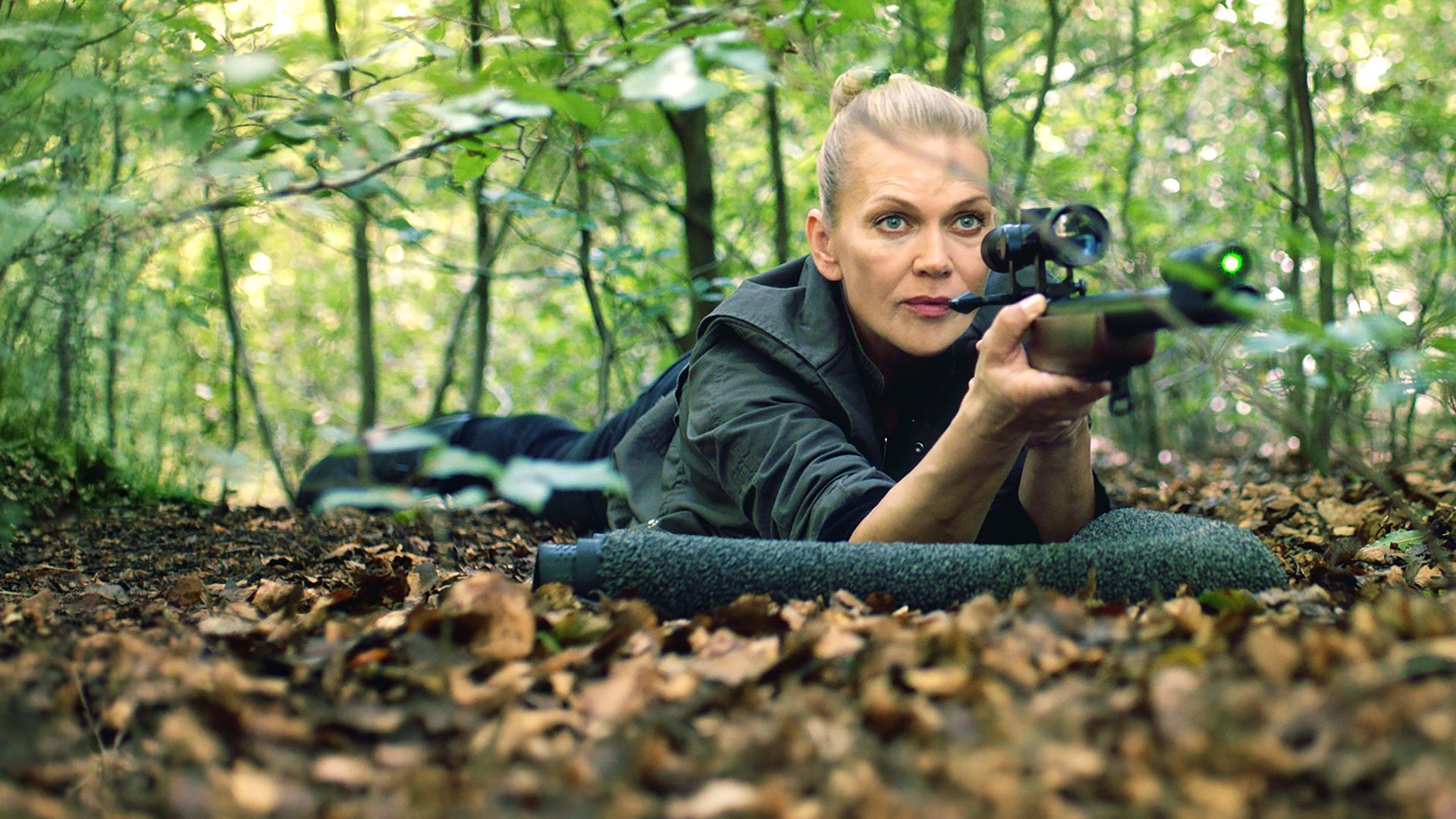 "Helen Dorn - Das Recht zu schweigen": Helen Dorn (Anna Loos) liegt bäuchlings mit einem Gewehr im Anschlag auf dem Waldboden.