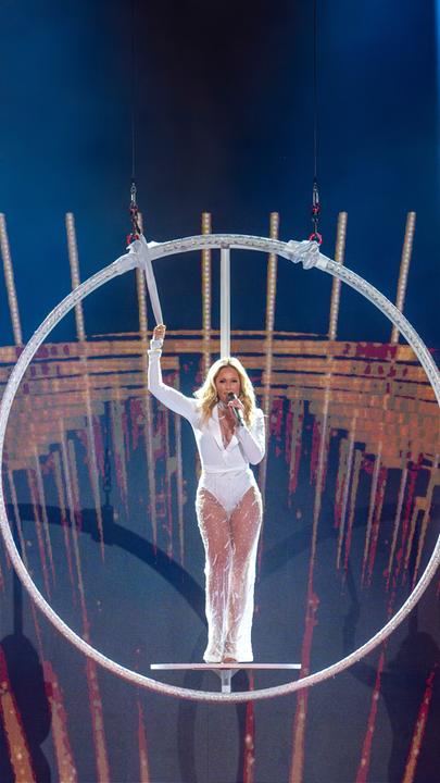 Helene Fischer in einem weißen Show-Outfit mit transparenten Hosenbeinen, in der Mitte einer silbernen Ringkonstruktion. Ihre rechte Hand ist mit einer Schlaufe am Ring gesichert, mit der linken Hand hält sie ein Mikrofon vor ihren Mund, in das sie singt. Sie schaut in Richtung Kamera. Das Bild zeigt eine totale Kameraeinstellung.
