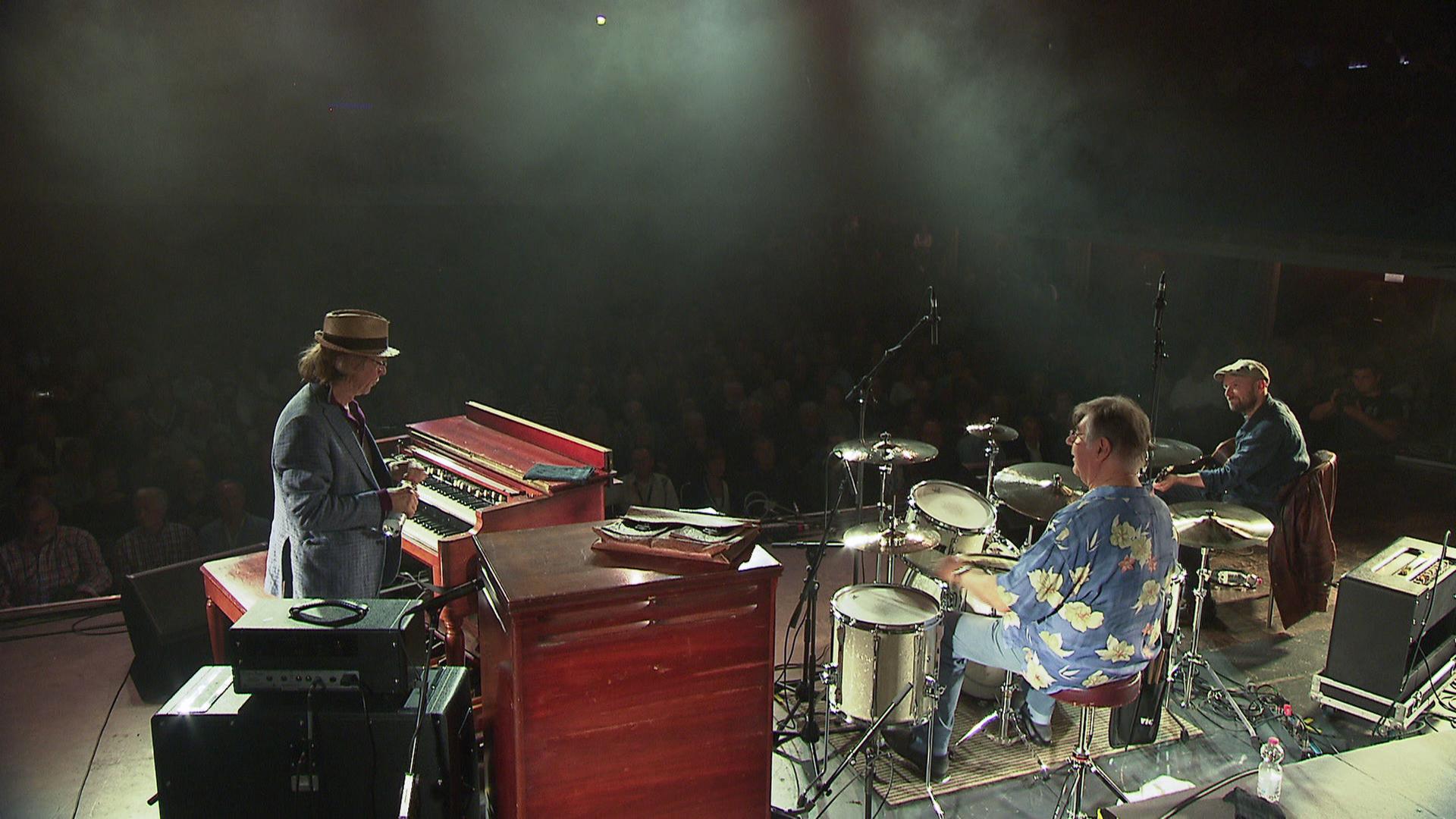 Foto einer großen, beleuchteten Bühne. Helge Schneider, Pete York und Gitarrist Henrik Freischlader sitzen an ihren Instrumenten. 