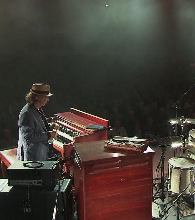 Foto einer großen, beleuchteten Bühne. Helge Schneider, Pete York und Gitarrist Henrik Freischlader sitzen an ihren Instrumenten. 