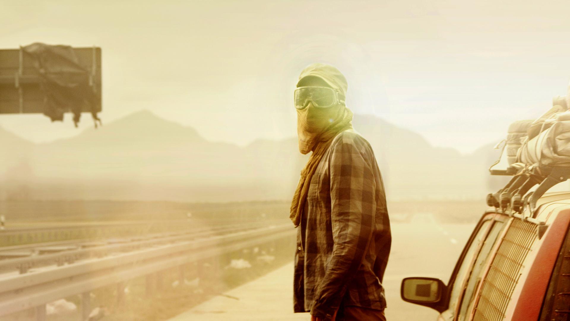 "Hell": Halbnahe Gegenlichtaufnahme mit leichtem Gelbfilter: Eine vermummte Person, mit Schildkappe, Skisonnenbrille und Tuch vor Mund, steht neben einem Auto auf einer verlassenen und zugemüllten Schnellstraße.