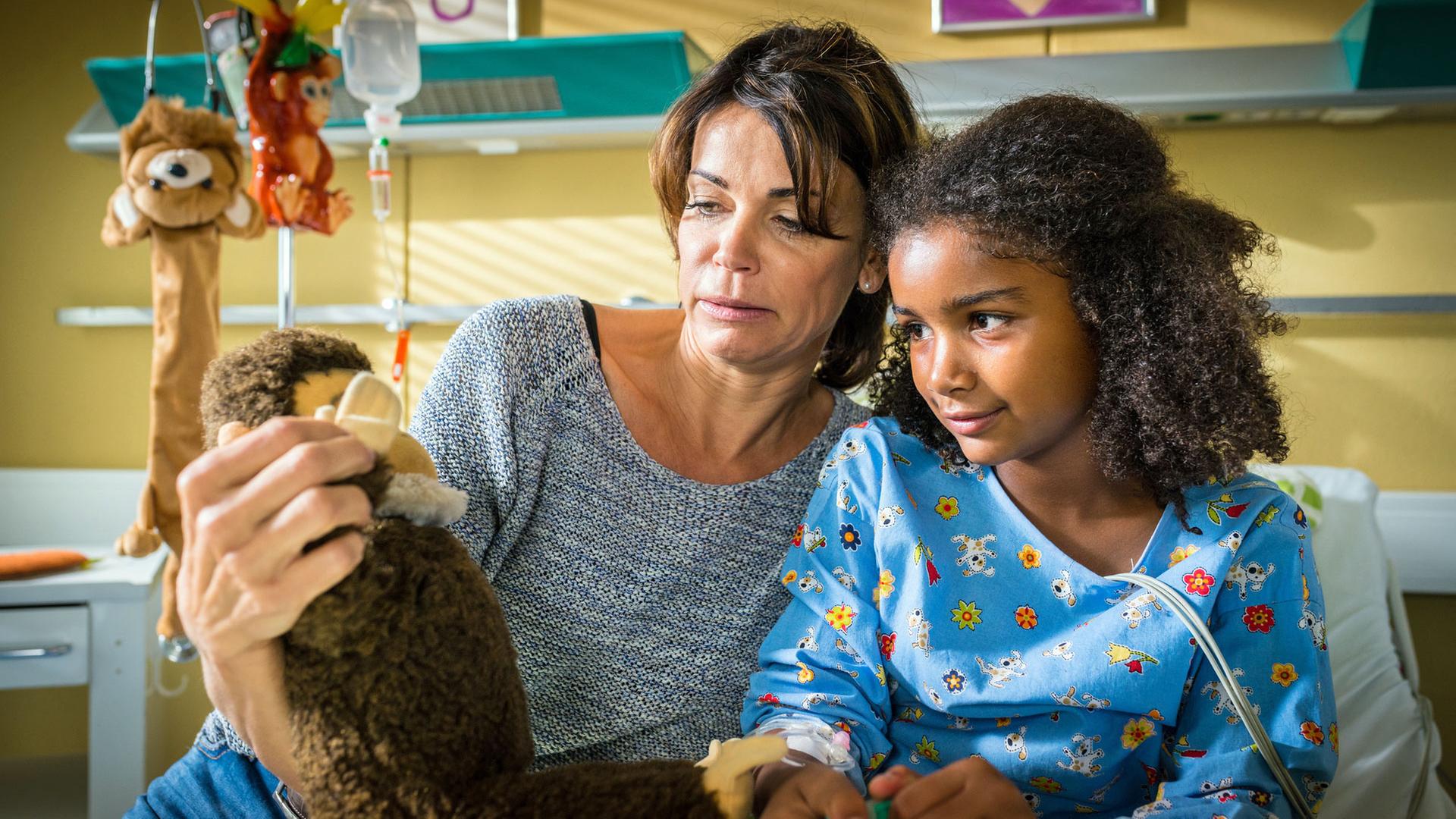 "Notruf Hafenkante - Help": Im Krankenzimmer sitzt Frau Dr. Jonas (Gerit Kling) mit ihrer kleinen Patientin (Rokhaya Boye) auf deren Krankenbett und zeigt ihr einen Plüschaffen.