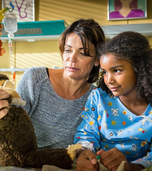 "Notruf Hafenkante - Help": Im Krankenzimmer sitzt Frau Dr. Jonas (Gerit Kling) mit ihrer kleinen Patientin (Rokhaya Boye) auf deren Krankenbett und zeigt ihr einen Plüschaffen.