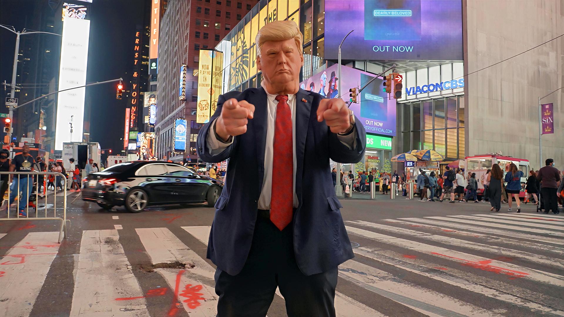 Ein Donald-Trump-Impersonator mit Maske steht nachts am Times Square in New York auf einem Zebrastreifen und zeigt mit beiden Händen in die Kamera.