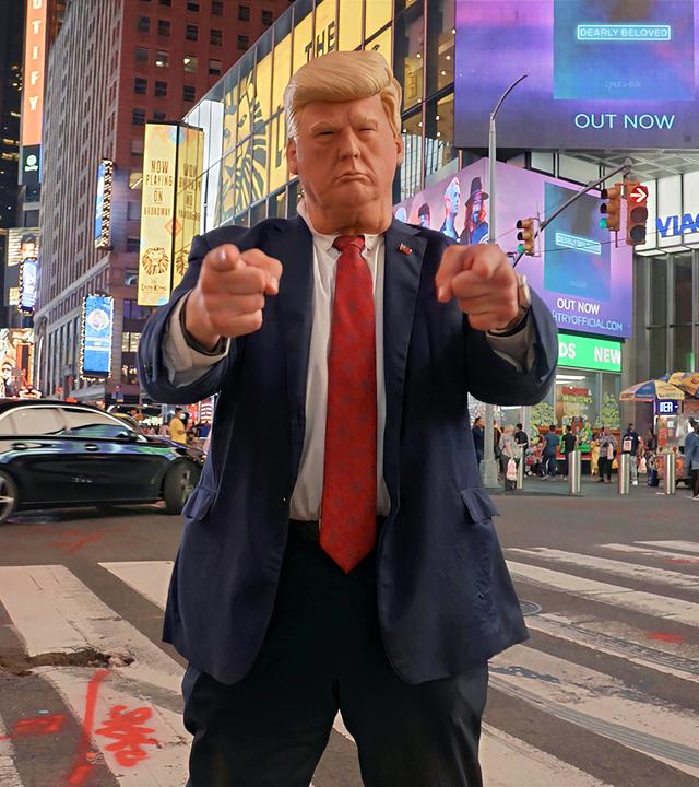 Ein Donald-Trump-Impersonator mit Maske steht nachts am Times Square in New York auf einem Zebrastreifen und zeigt mit beiden Händen in die Kamera.