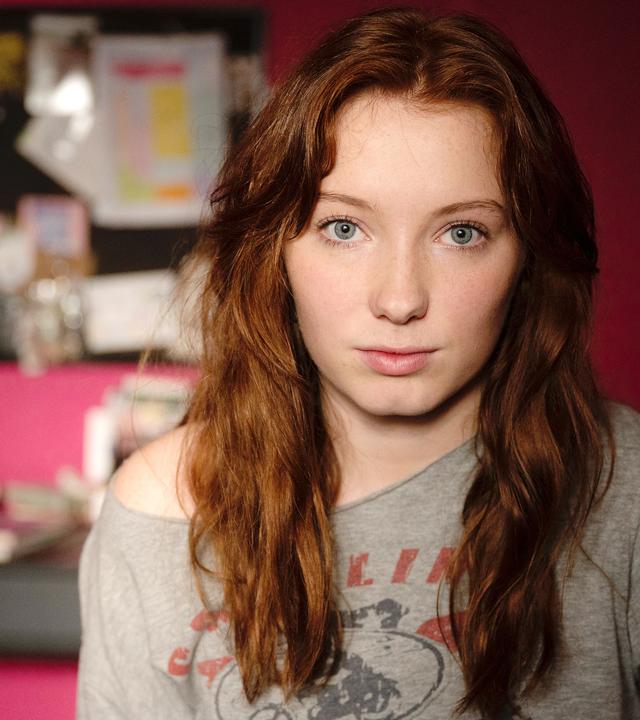 Joanna (Nikki Hanseblad) steht in einem Zimmer vor einer roten Wand.