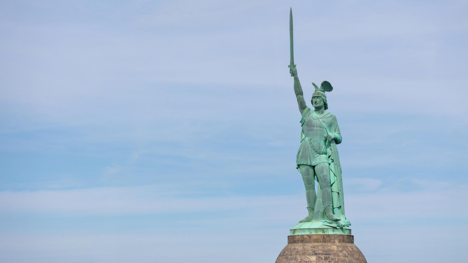 Mit grüner Patina überzogene Statue eines germanischen Kriegers, der einen geflügelten Helm trägt und mit der rechten Hand ein Schwert in den Himmel reckt.