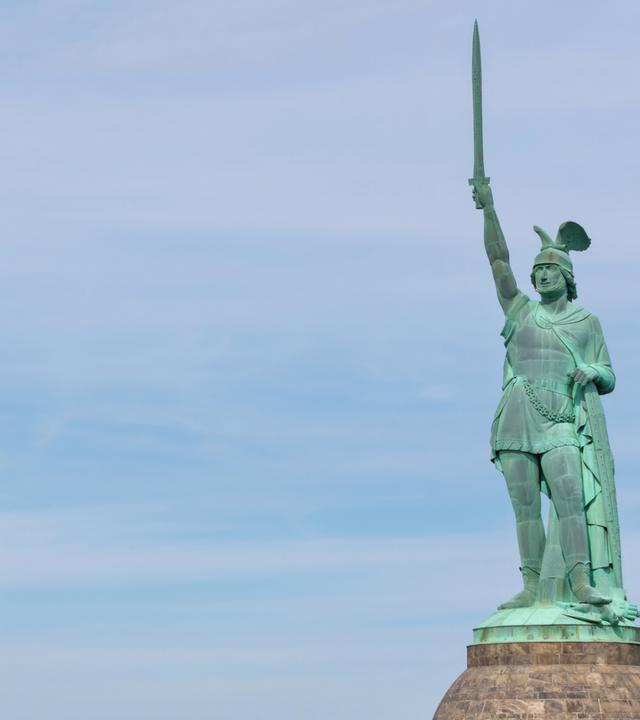 Mit grüner Patina überzogene Statue eines germanischen Kriegers, der einen geflügelten Helm trägt und mit der rechten Hand ein Schwert in den Himmel reckt.