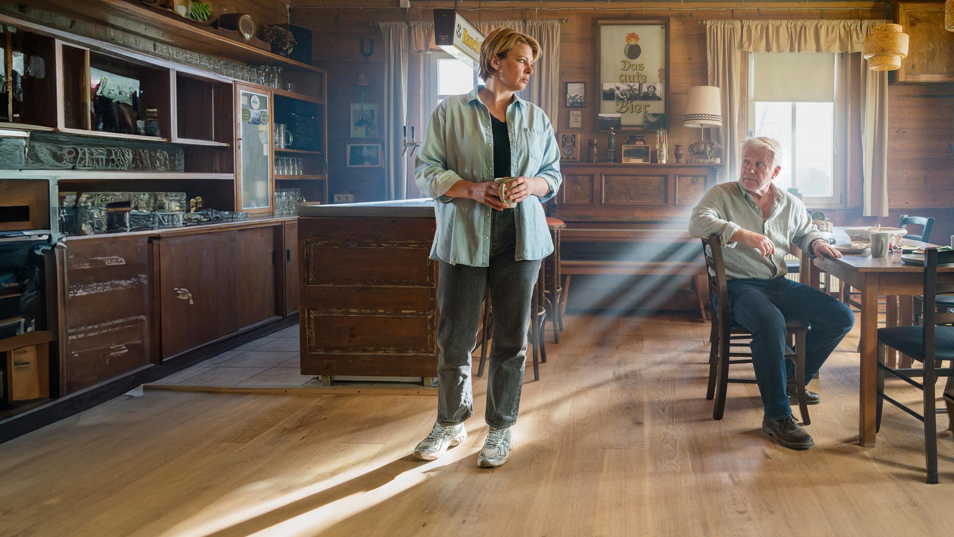 Ina Sedlitz (Jördis Triebel) steht in einem rustikal eingerichteten Gastraum aus Holz und blickt zu Walter Sedlitz (Jörg Schüttauf), der an einem Tisch sitzt. 