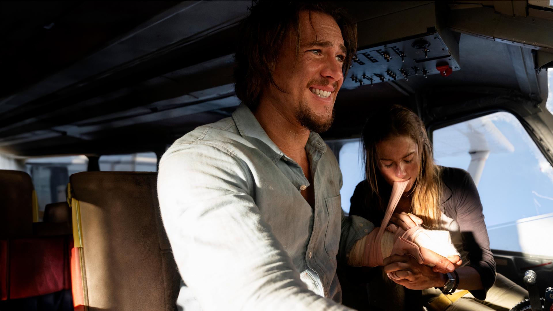 Sara (Allison Williams) und Jackson (Alexander Dreymon) sitzen im Cockpit eines Kleinflugzeuges. Sara legt Jackson einen Druckverband an und hält den Stoff mit ihren Zähnen stramm. Er hat sichtbare Schmerzen und schaut weg.