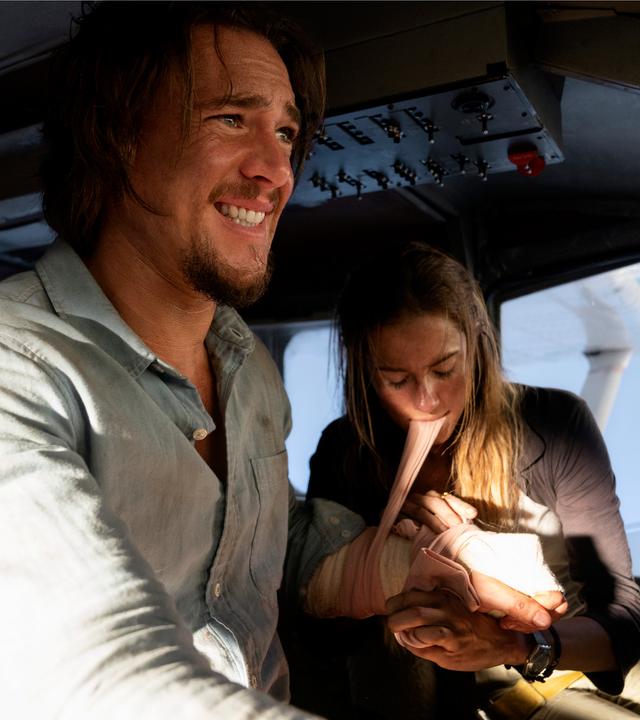 Sara (Allison Williams) und Jackson (Alexander Dreymon) sitzen im Cockpit eines Kleinflugzeuges. Sara legt Jackson einen Druckverband an und hält den Stoff mit ihren Zähnen stramm. Er hat sichtbare Schmerzen und schaut weg.
