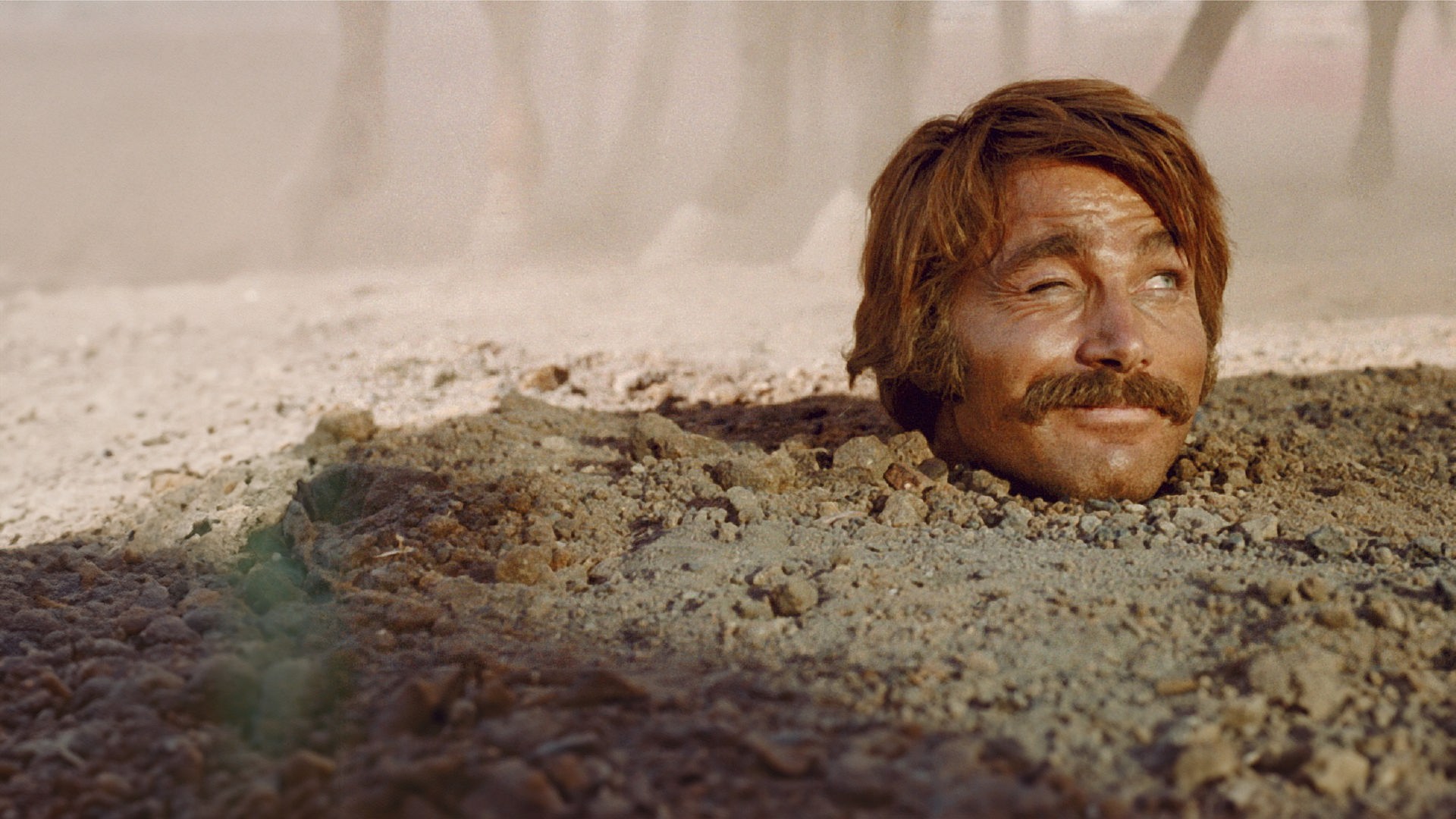 Yodlaf Peterson (Franco Nero) steckt bis zum Hals im Sand und blickt zwinkernd zur Seite. Im Hintergrund sind Pferdebeine in staubiger Luft zu erkennen.