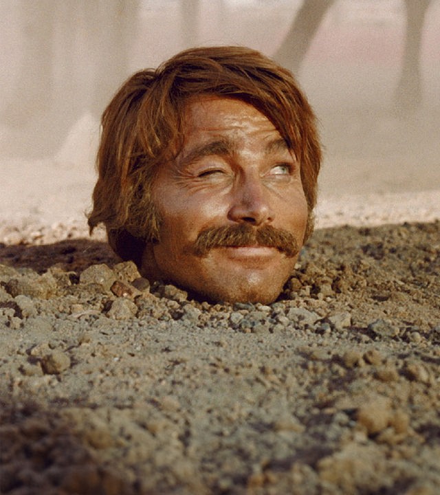Yodlaf Peterson (Franco Nero) steckt bis zum Hals im Sand und blickt zwinkernd zur Seite. Im Hintergrund sind Pferdebeine in staubiger Luft zu erkennen.