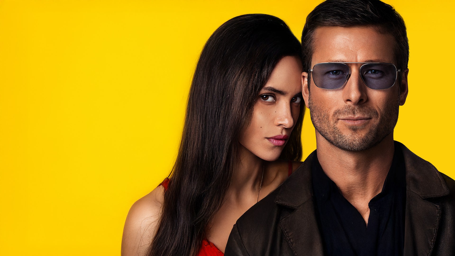 Nahaufnahme: Vor einem gelben Hintergrund schaut Gary (Glen Powell) mit Dreitagebart und Sonnenbrille nach vorne. Seitlich von ihm blickt Madison (Adria Arjona) über seine Schulter.