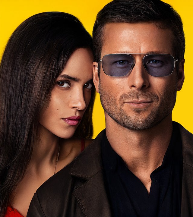 Nahaufnahme: Vor einem gelben Hintergrund schaut Gary (Glen Powell) mit Dreitagebart und Sonnenbrille nach vorne. Seitlich von ihm blickt Madison (Adria Arjona) über seine Schulter.