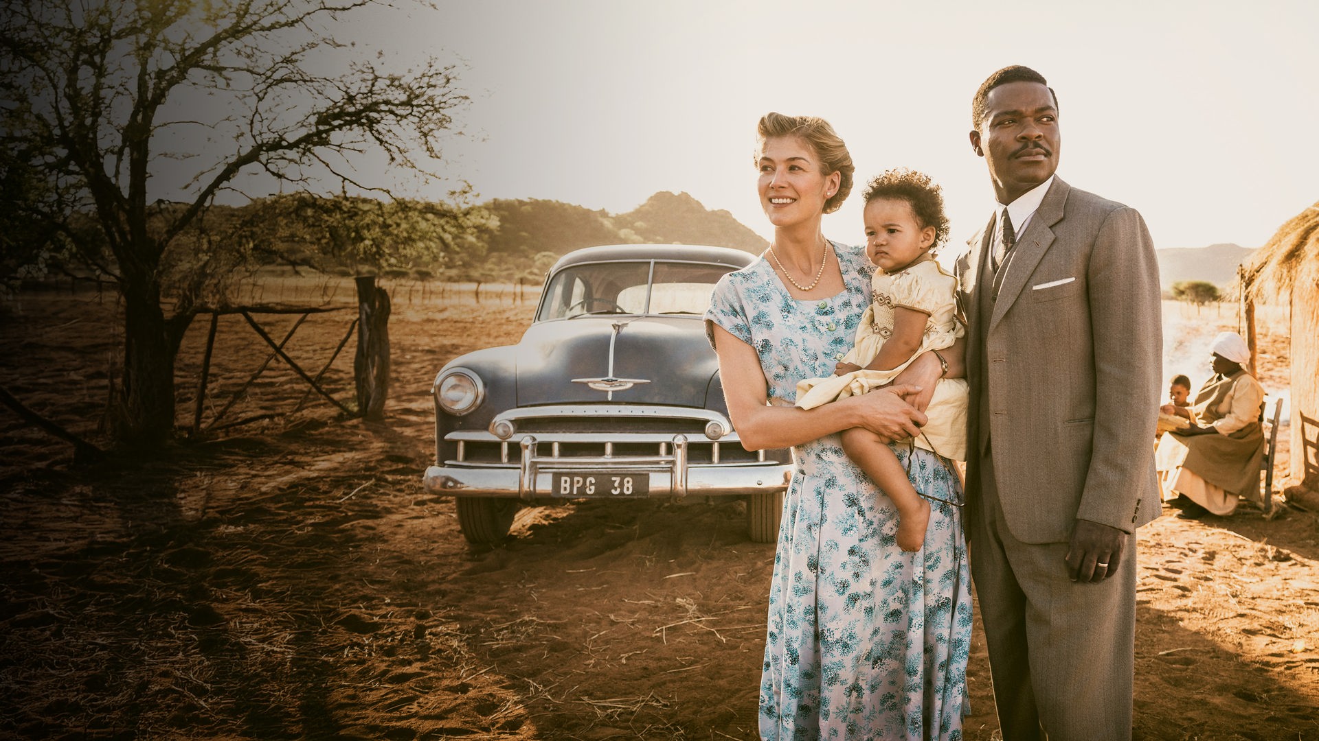 "A United Kingdom": Vor einem afrikanischen Dorf: Ruth Williams (Rosamund Pike) trägt ihre kleine Tochter auf dem Arm und steht neben Seretse Khama (David Oyelowo). Im Hintergrund steht ein Auto vor einer Hütte.