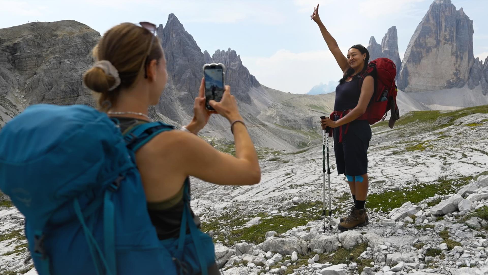In einer felsigen Hochgebirgslandschaft fotografiert Nina ihre Begleiterin Djenna mit dem Smartphone. Djenna trägt einen großen roten Wanderrucksack, hält zwei Wanderstöcke und hebt lächelnd einen Arm. Im Hintergrund ragen mehrere markante graue Felsgipfel in den hellen Himmel.