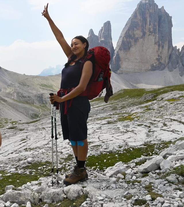 In einer felsigen Hochgebirgslandschaft fotografiert Nina ihre Begleiterin Djenna mit dem Smartphone. Djenna trägt einen großen roten Wanderrucksack, hält zwei Wanderstöcke und hebt lächelnd einen Arm. Im Hintergrund ragen mehrere markante graue Felsgipfel in den hellen Himmel.