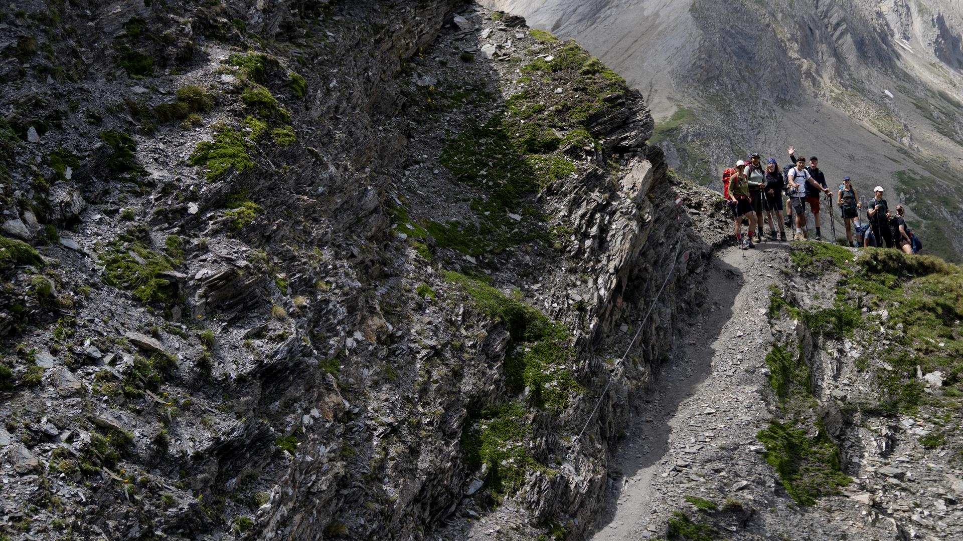 Wanderer mit Rucksäcken steigen hintereinander über einen schmalen, felsigen Bergpfad. Die Wanderin im Vordergrund beugt sich beim steilen Abstieg nach vorn, blickt konzentriert auf den Boden und sucht Halt am schmalen Drahteil.