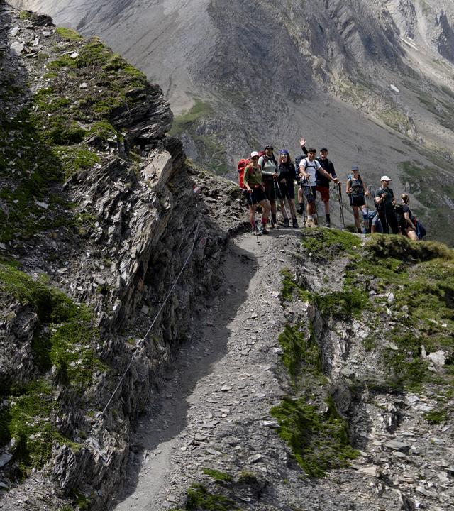 Wanderer mit Rucksäcken steigen hintereinander über einen schmalen, felsigen Bergpfad. Die Wanderin im Vordergrund beugt sich beim steilen Abstieg nach vorn, blickt konzentriert auf den Boden und sucht Halt am schmalen Drahteil.