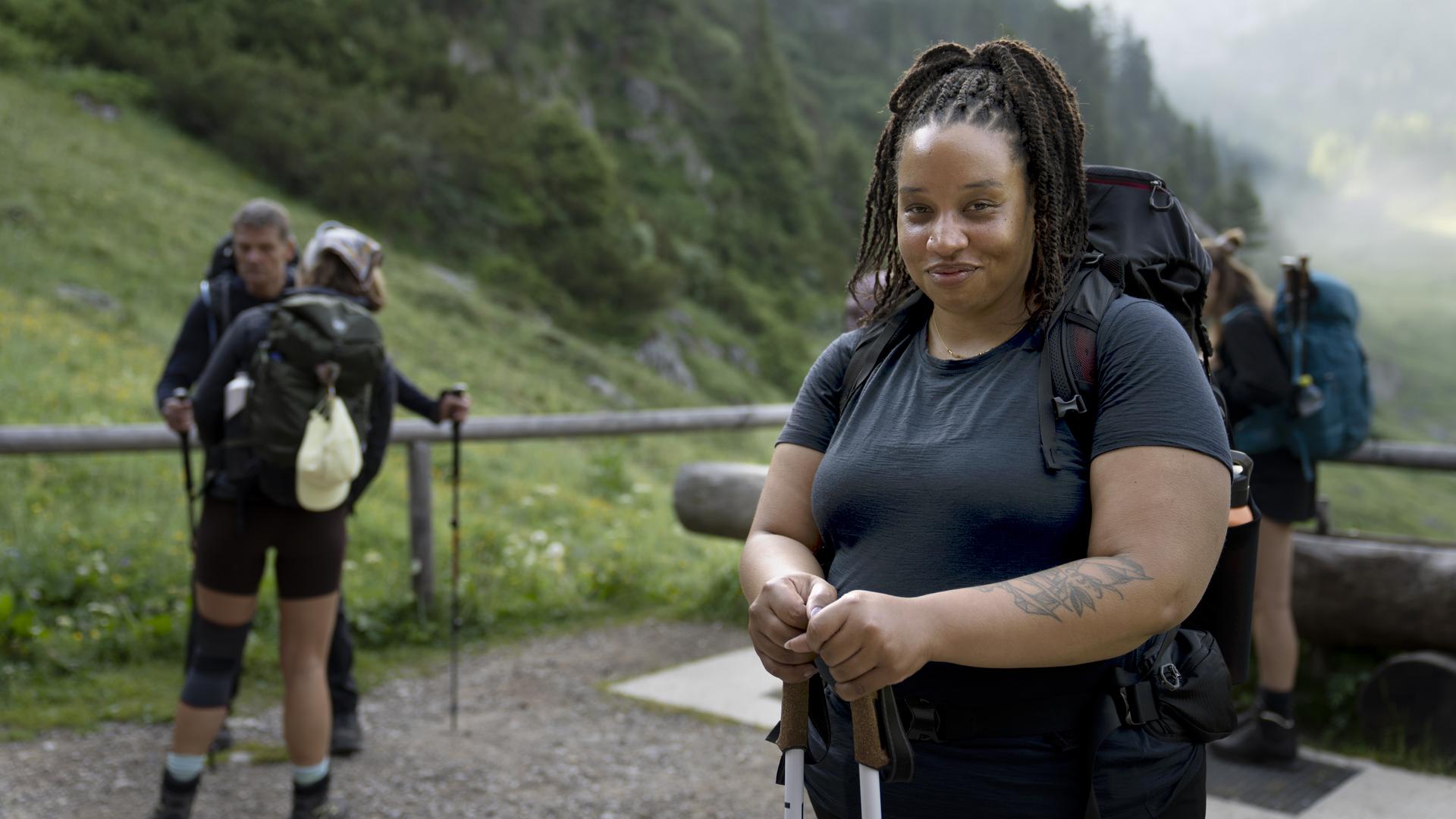 Im Vordergrund lächelt eine Wanderin, während sie Wanderstöcke hält. Ihr großer Rucksack und die sportliche Kleidung weisen auf eine längere Tour hin. Hinter ihr stehen mehrere Wandernde auf einem schmalen Weg inmitten einer grünen Berglandschaft.