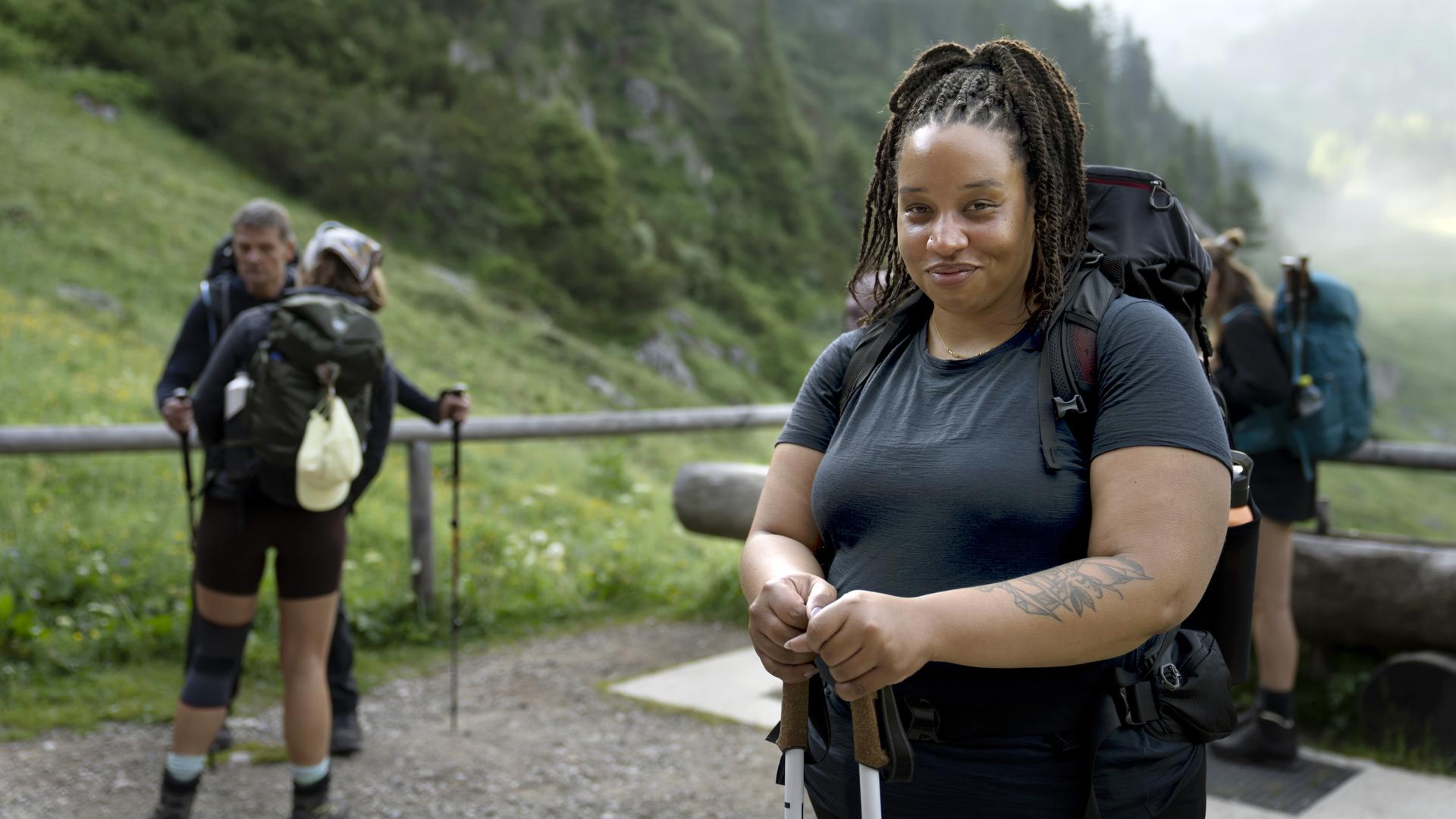 Im Vordergrund lächelt eine Wanderin, während sie Wanderstöcke hält. Ihr großer Rucksack und die sportliche Kleidung weisen auf eine längere Tour hin. Hinter ihr stehen mehrere Wandernde auf einem schmalen Weg inmitten einer grünen Berglandschaft.