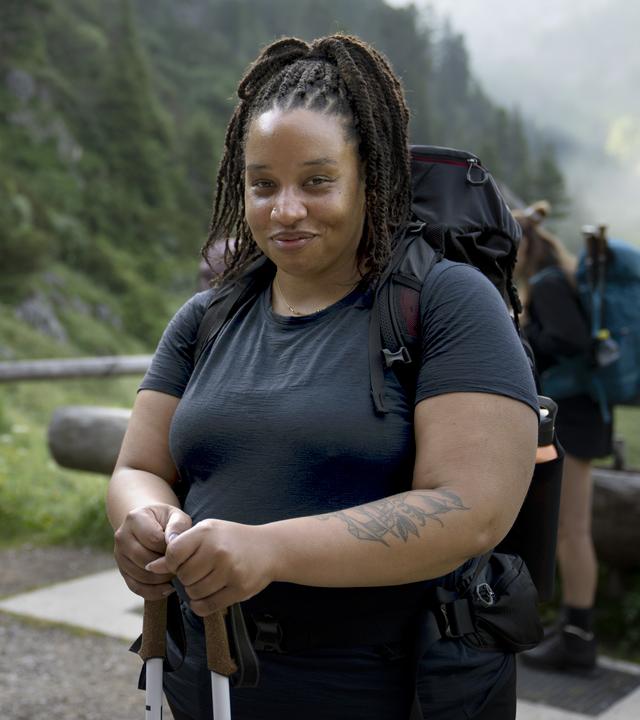 Im Vordergrund lächelt eine Wanderin, während sie Wanderstöcke hält. Ihr großer Rucksack und die sportliche Kleidung weisen auf eine längere Tour hin. Hinter ihr stehen mehrere Wandernde auf einem schmalen Weg inmitten einer grünen Berglandschaft.
