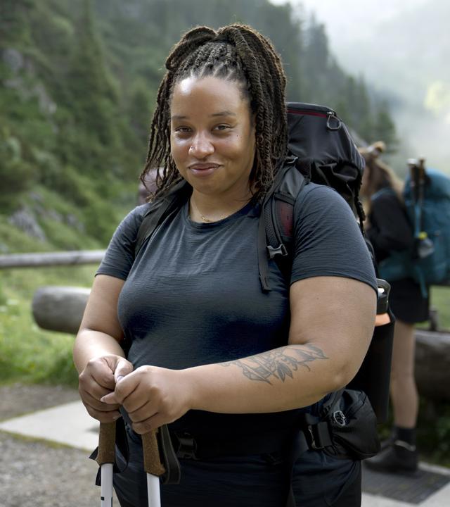 Im Vordergrund lächelt eine Wanderin, während sie Wanderstöcke hält. Ihr großer Rucksack und die sportliche Kleidung weisen auf eine längere Tour hin. Hinter ihr stehen mehrere Wandernde auf einem schmalen Weg inmitten einer grünen Berglandschaft.
