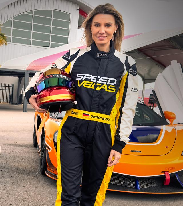 Eine blonde Frau posiert, in einen schwarz-gelben Rennfahr-Anzug gekleidet, zwischen mehreren geparkten Rennautos. Sie hat einen Helm mit Visier unterm Arm und lächelt in die Kamera.