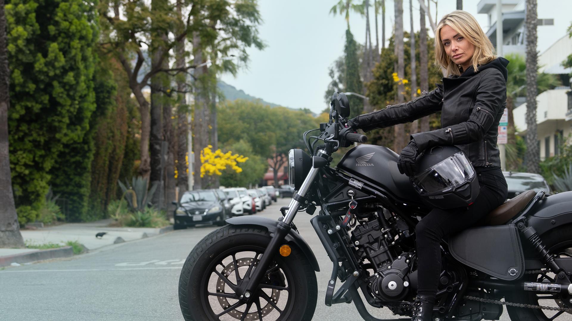 Eine junge blonde Frau sitzt, gekleidet in schwarzer Motorradjacke, auf einem schwarzen Motorrad. Das Motorrad steht auf einer Straßenkreuzung in Beverly Hills mit Palmen im Hintergrund.