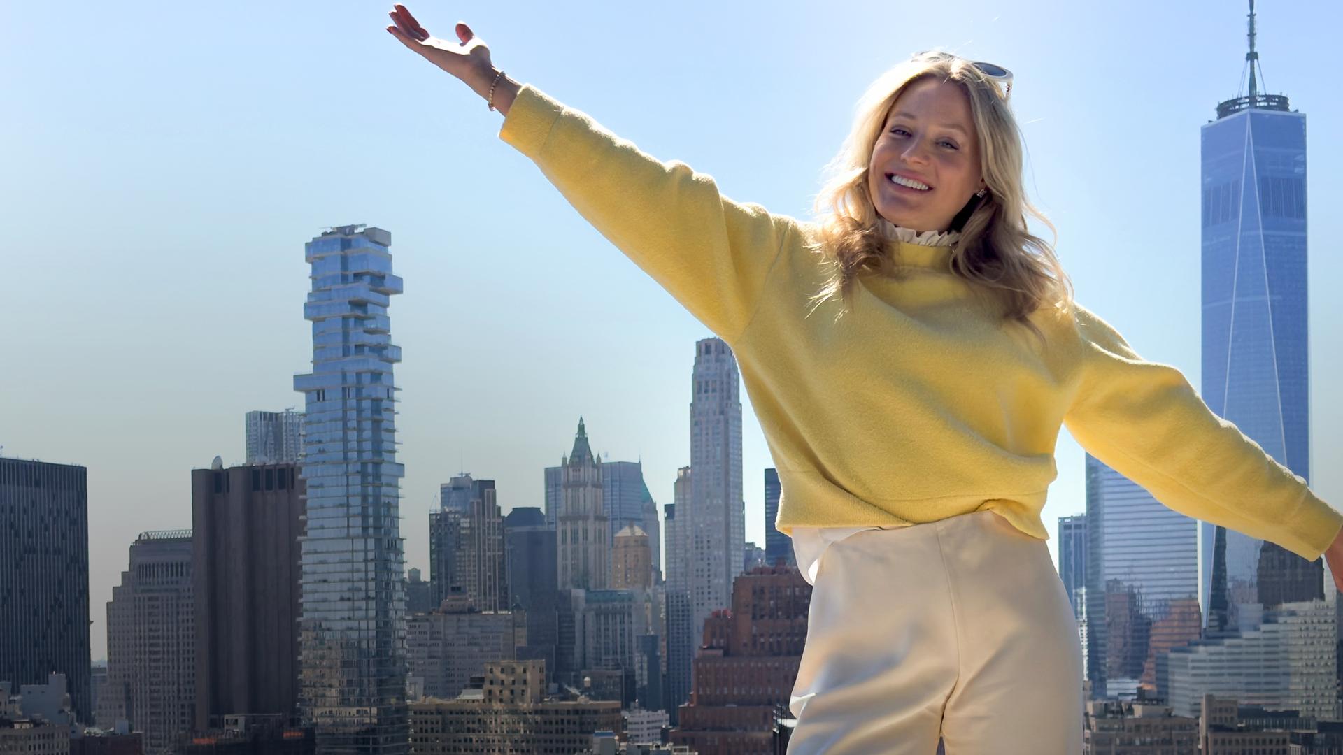 Eine 30 Jahre alte Frau steht, in gelbem Pulli gekleidet, mit ausgestreckten Armen, auf dem Dach eines Hochhauses in New York. Lächelnd blickt sie in die Kamera, während im Hintergrund die Skyline von New York in der Sonne glänzt.