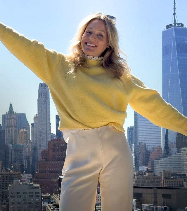 Eine 30 Jahre alte Frau steht, in gelbem Pulli gekleidet, mit ausgestreckten Armen, auf dem Dach eines Hochhauses in New York. Lächelnd blickt sie in die Kamera, während im Hintergrund die Skyline von New York in der Sonne glänzt.