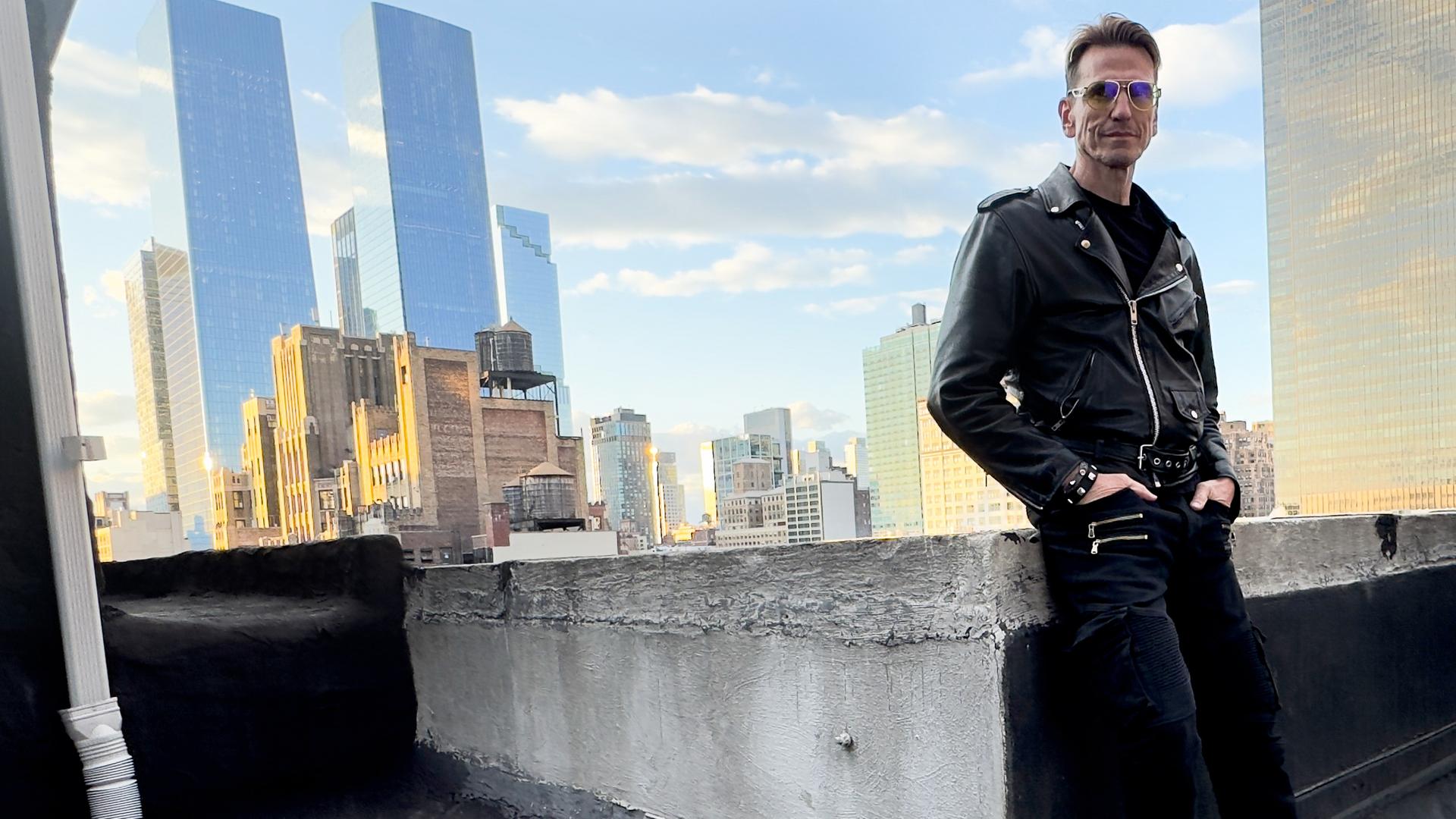 Ein circa 50 Jahre alter Mann steht zwischen Gebäuden und hält eine Fotokamera mit großem Objektiv in der Hand. In ist in eine dunkle Jacke gekleidet und trägt eine gelbe, auffällige Brille. Die Sonne blitzt zwischen den Gebäuden über seiner Schulter hervor.