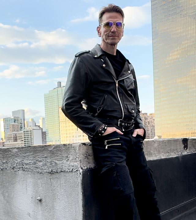 Ein circa 50 Jahre alter Mann steht zwischen Gebäuden und hält eine Fotokamera mit großem Objektiv in der Hand. In ist in eine dunkle Jacke gekleidet und trägt eine gelbe, auffällige Brille. Die Sonne blitzt zwischen den Gebäuden über seiner Schulter hervor.