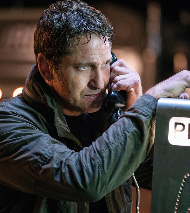 "Angel Has Fallen": Mike Banning (Gerard Butler) steht verschwitzt vor einem öffentlichen Telefon, auf das er seinen rechten Arm leicht abstützt und mit der anderen Hand den Hörer zum Telefonieren hält.