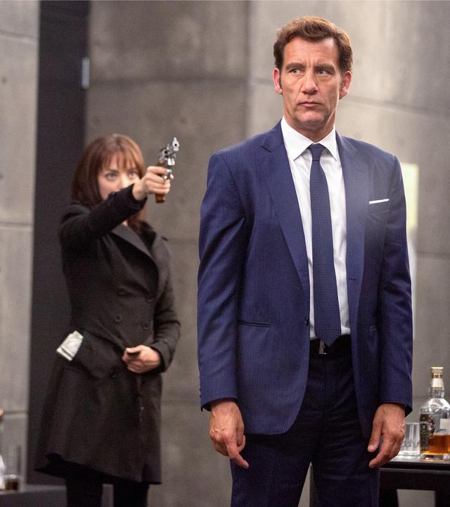 Sal (Clive Owen) steht in einem großen Raum neben dem Tisch. Hinter ihm steht Anon (Amanda Seyfried) und zielt mit der Pistole auf ihn.