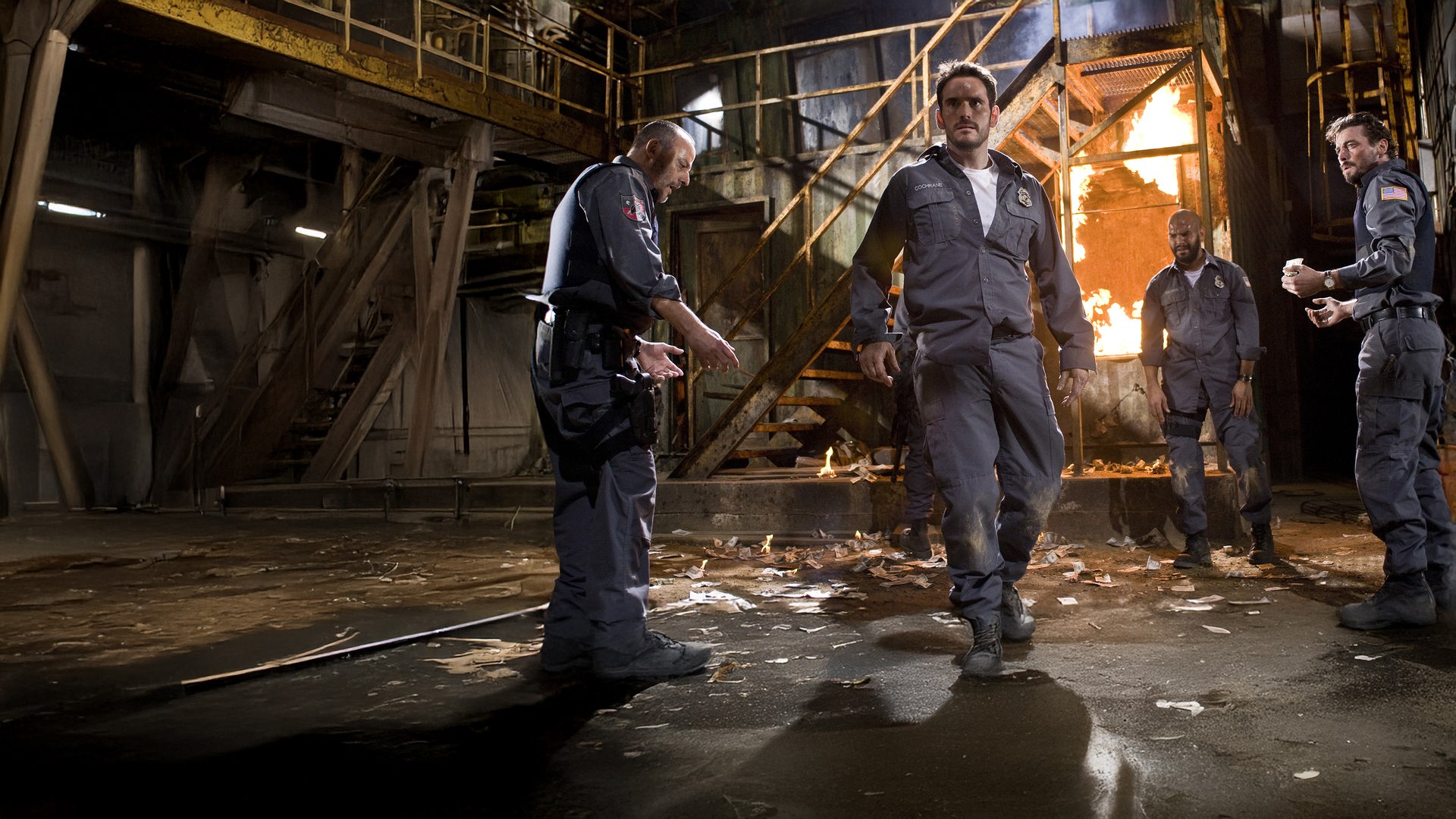 "Armored": Die uniformierten Wächter eines Geldtransports Quinn (Jean Reno), Mike Cochrane (Matt Dillon), Palmer (Amaury Nolasco) und Dobbs (Skeet Ulrich) stehen in einer Fabrikhalle vor einer brennenden Treppe und schauen angespannt zur anderen Seite des Raumes.